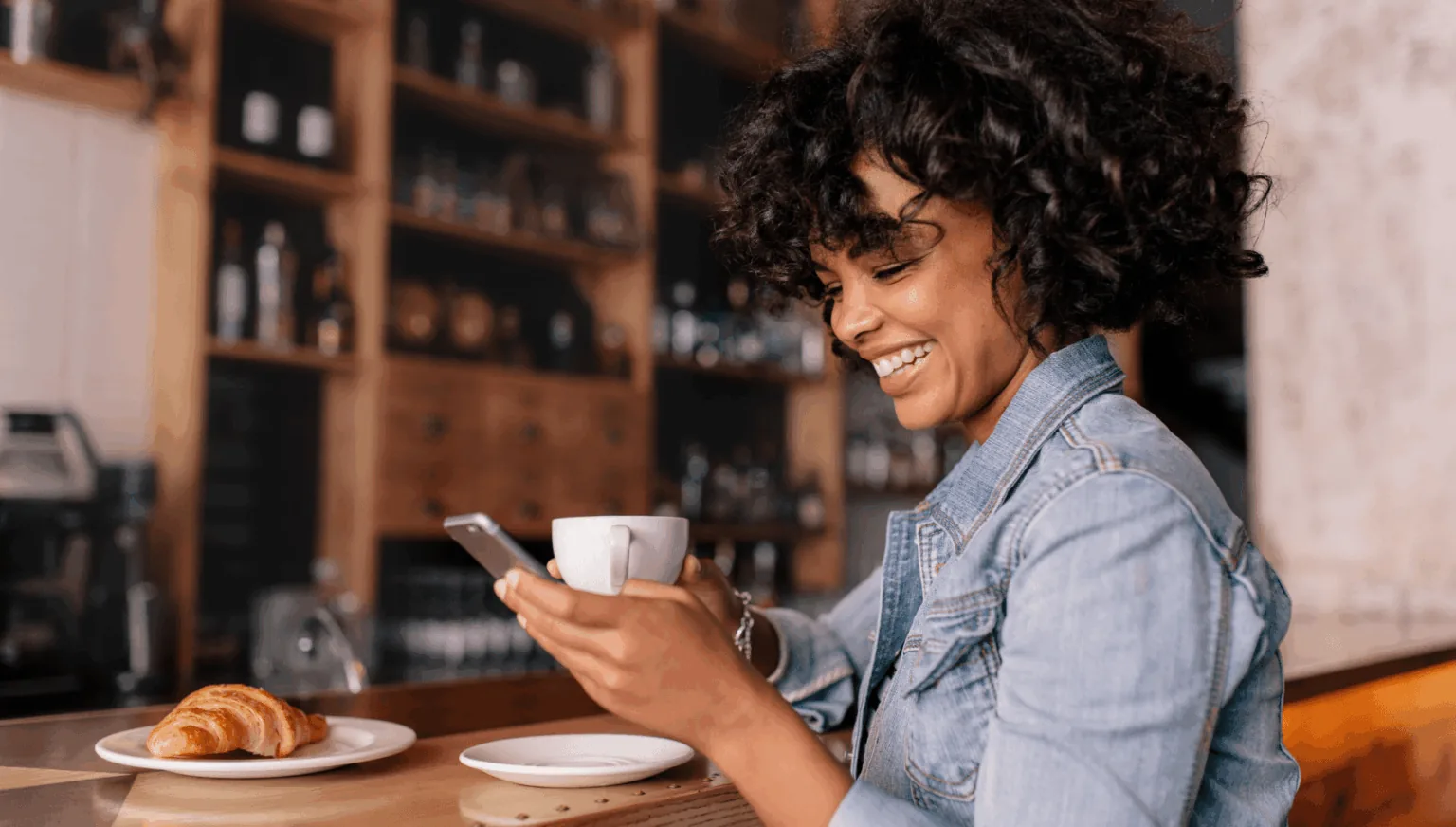 Woman smiling at her phone while holding a cup of coffee in a café
