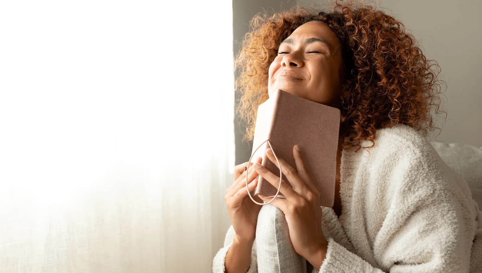 Woman with curly hair smiling with her eyes closed, hugging a notebook near a sunny window